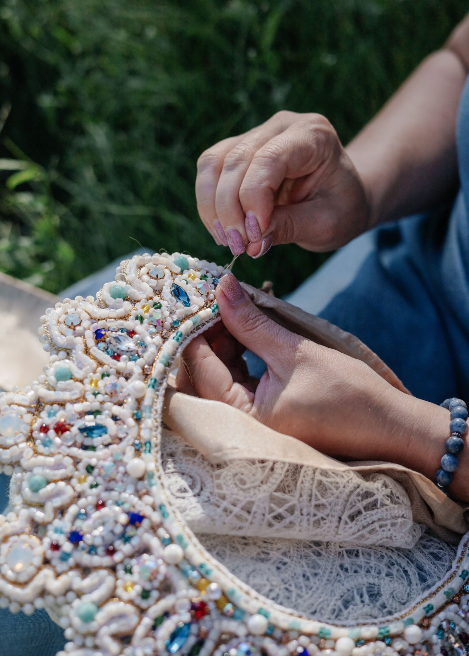 Close-up of hands crafting detailed bead embroidery on fabric outdoors.